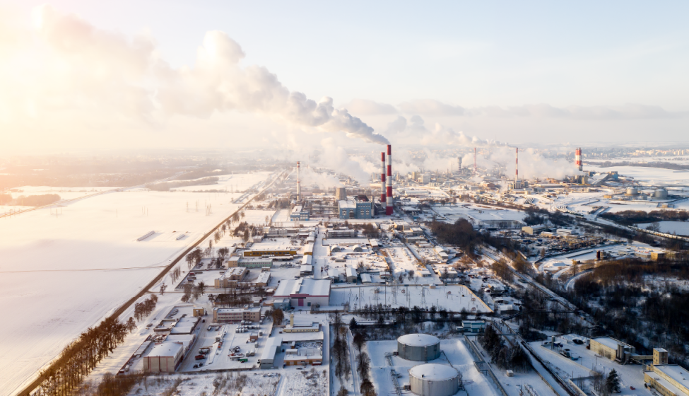 Aerial view of snow-covered industrial section of city with red and white striped smokestacks blowing smoke across hazy, dim-lit sky