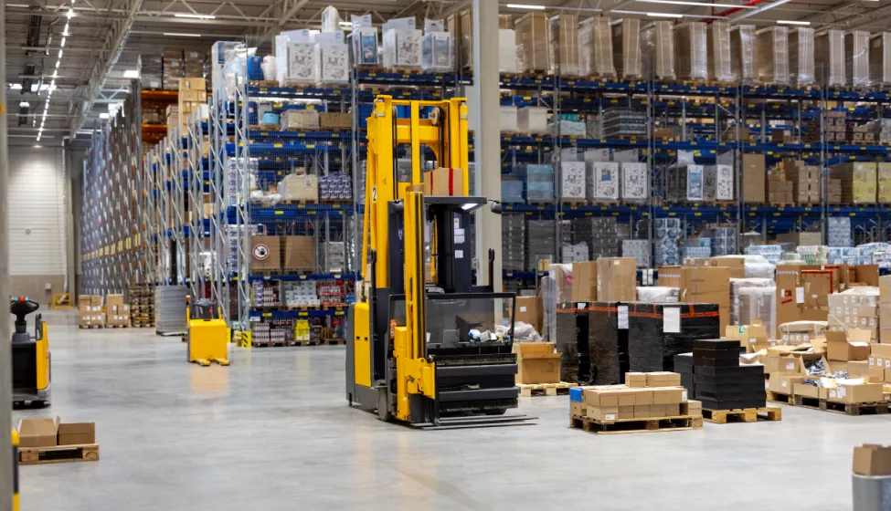 A forklift sits empty inside a product warehouse surrounded by pallets and boxes with stacked shelves in the background