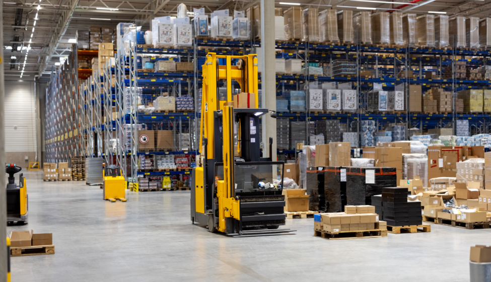 A forklift sits empty inside a product warehouse surrounded by pallets and boxes with stacked shelves in the background