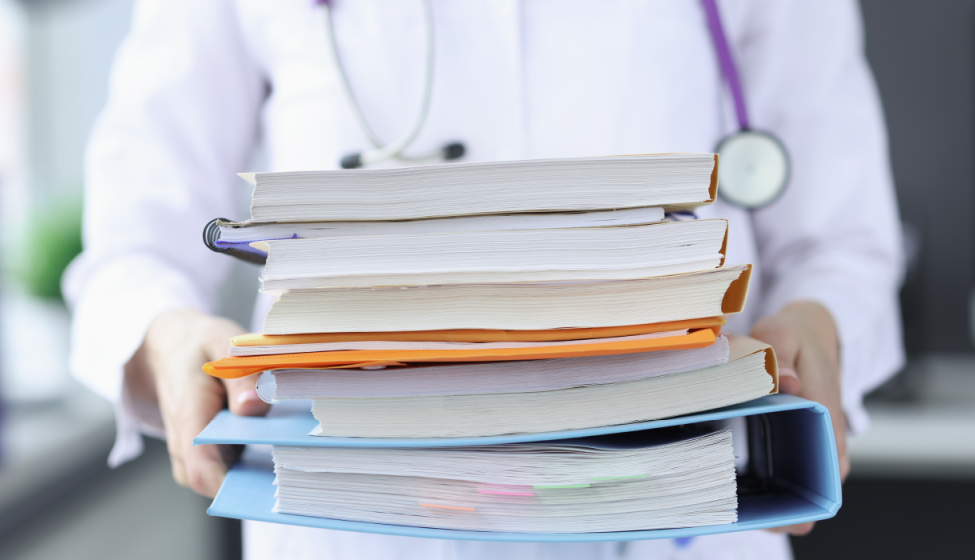 A healthcare worker holds a large stack of paperwork and folders while wearing a stethoscope around their neck
