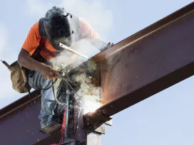 A worker is elevated above a building welding an I-beam. Exponent materials engineers consult on welding and fabrication. 