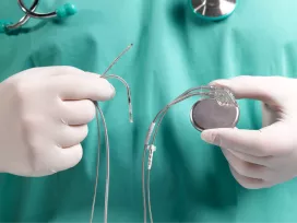 Gloved, medical professional in green scrubs with stethoscope around neck (shown from neck to mid torso) holding an implanted medical device